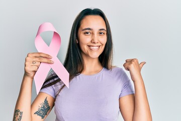 Young hispanic woman holding pink cancer ribbon pointing thumb up to the side smiling happy with open mouth