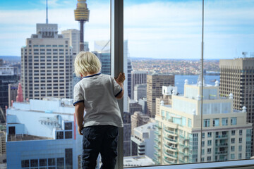 Little boy standing at window of high rise apartment looking down at view of the city below