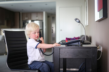 Little boy sitting at desk pretending to talk on phone in bright apartment