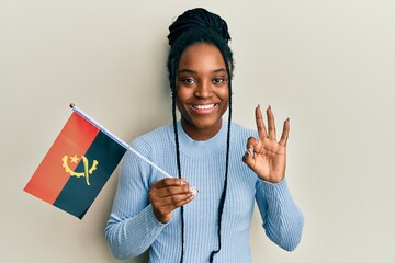 African american woman with braided hair holding angola flag doing ok sign with fingers, smiling friendly gesturing excellent symbol - obrazy, fototapety, plakaty