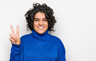 Young hispanic woman with curly hair wearing turtleneck sweater showing and pointing up with fingers number two while smiling confident and happy.