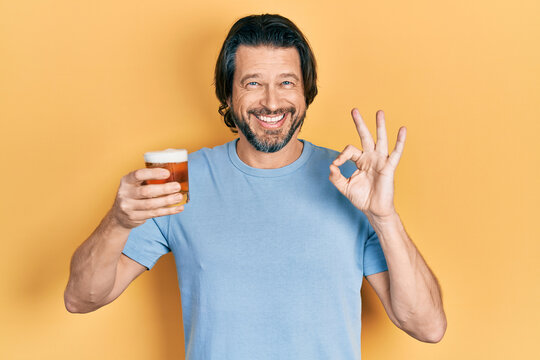 Middle age caucasian man drinking a pint of beer doing ok sign with fingers, smiling friendly gesturing excellent symbol
