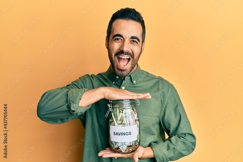 Poster Young hispanic man holding jar with savings smiling and laughing hard out loud because funny crazy joke.