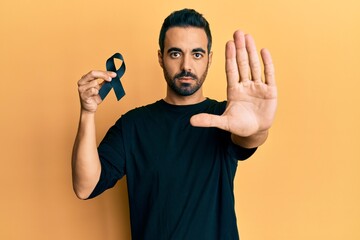Young hispanic man holding black ribbon with open hand doing stop sign with serious and confident...