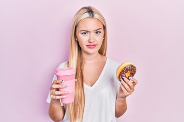Young blonde girl eating doughnut and drinking coffee skeptic and nervous, frowning upset because...