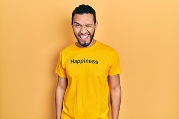 Hispanic man with beard wearing t shirt with happiness word message winking looking at the camera with sexy expression, cheerful and happy face.
