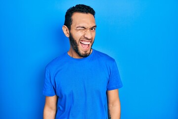 Hispanic man with beard wearing casual blue t shirt winking looking at the camera with sexy expression, cheerful and happy face.