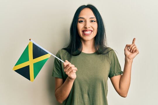 Young hispanic girl holding jamaica flag smiling happy pointing with hand and finger to the side