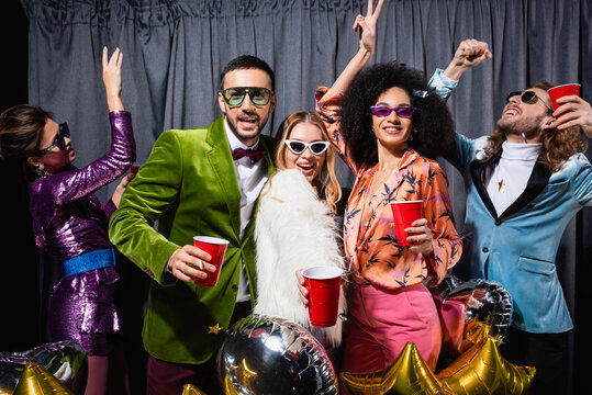 positive interracial friends in colorful clothes and sunglasses dancing near grey curtain on black background.