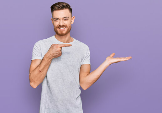 Young redhead man wearing casual grey t shirt amazed and smiling to the camera while presenting with hand and pointing with finger.