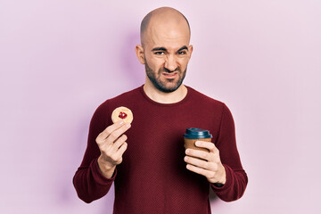 Young bald man drinking coffee and eating pastry clueless and confused expression. doubt concept.