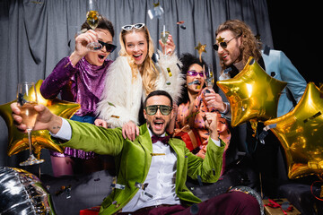 positive interracial friends in sunglasses and colorful clothes drinking champagne near grey curtain on black background.