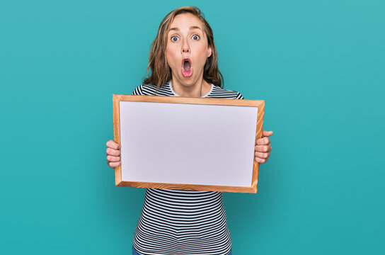 Young blonde woman holding empty white board scared and amazed with open mouth for surprise, disbelief face