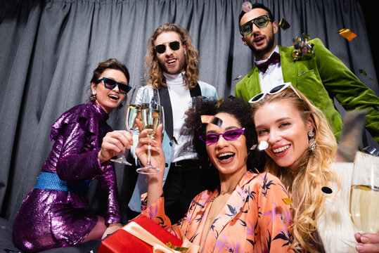 smiling interracial friends in sunglasses and colorful clothes drinking champagne on grey background.