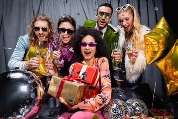 happy african american woman with gift boxes sitting among interracial friends near grey curtain on black background.