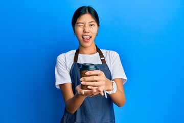 Young chinese woman wearing waiter apron holding cup of coffee winking looking at the camera with sexy expression, cheerful and happy face.