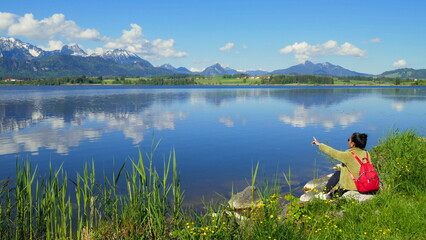 junge Frau genießt die stille Natur am Hopfensee im Allgäu am grünen Ufer sitzend bei blauem Himmel 