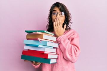 Young hispanic woman holding a pile of books covering mouth with hand, shocked and afraid for mistake. surprised expression