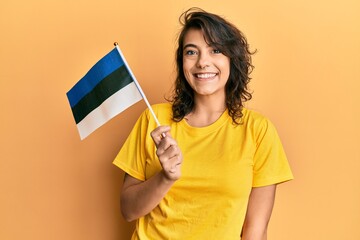 Young hispanic woman holding estonia flag looking positive and happy standing and smiling with a confident smile showing teeth