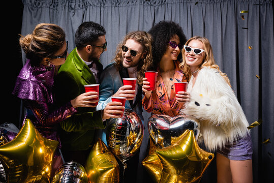 happy interracial friends in sunglasses celebrating with plastic cups near grey curtain on black background.