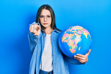 Young hispanic girl holding world ball pointing with finger to the camera and to you, confident gesture looking serious