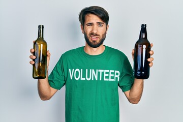Young hispanic man wearing volunteer t shirt holding recycling bottle glass clueless and confused...