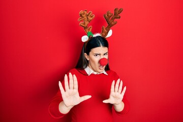 Young hispanic woman wearing deer christmas hat and red nose moving away hands palms showing...