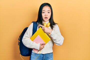 Young chinese girl holding student backpack and books surprised pointing with finger to the side, open mouth amazed expression.