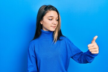 Young brunette girl wearing turtleneck sweater looking proud, smiling doing thumbs up gesture to the side