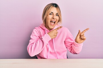 Young caucasian woman wearing casual clothes sitting on the table smiling and looking at the camera pointing with two hands and fingers to the side.