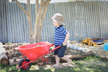 Little toddler boy pushing kid's wheelbarrow. Backyard sandpit play.