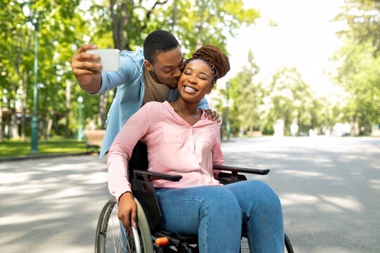 Romantic black man and his impaired girlfriend in wheelchair taking selfie together, kissing at city park