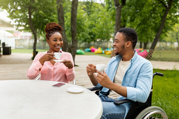 Disabled young black guy in wheelchair on date with pretty woman in outdoor cafe, drinking coffee,...
