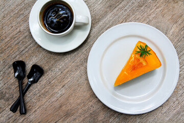 Top view piece of orange cake in white dish or plate, white cup of dark coffee and spoon on wooden background with copy space. Flat lay of sweet or dessert food on wood table. Snack set for eating.