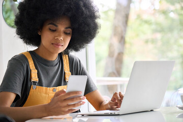 Young generation z black college student girl with afro hair sitting at table in cafe using mobile shop marketing application online with computer working texting surfing internet. - obrazy, fototapety, plakaty