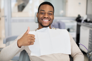 Happy young black man sitting in dentist chair and showing thumb up