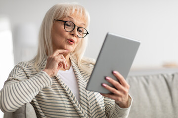 Surprised mature woman using tablet sitting on couch