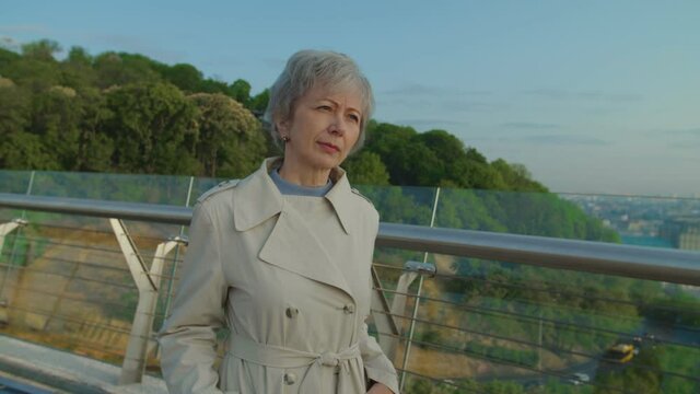 Senior woman in casual clothing standing in city park waiting for husband. Elderly bearded man approaching woman and presenting flowers. She expressing exitement and surprising to meet her spouse