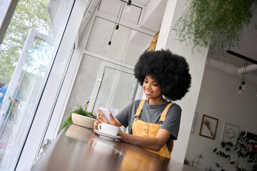 Young happy gen z black student hipster girl with afro hair sitting at table in cafe indoor using mobile shop marketing app online watching video working typing surfing internet. - obrazy, fototapety, plakaty