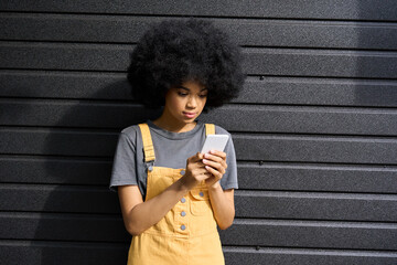 Fashion blogger young stylish afro American 20s girl holding smartphone standing on black wall...