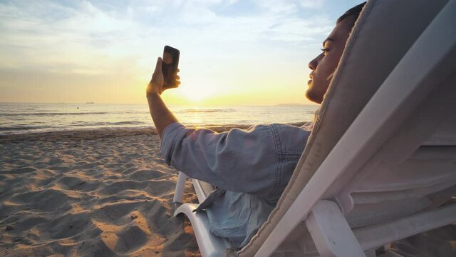 Young enjoying on the lounge chair on the sany tropical beach at sunset, sunrise and taking a selfie with smart phone, vacation and summer travel