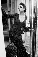 Young fashionable brunette lady with hairstyle in long black dress with glittering diamonds posing sad in fitting room with large mirrors - 437727419