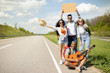 Joyful multiethnic friends thumbing their ride on highway with empty sign, playing guitar, going on autostop journey