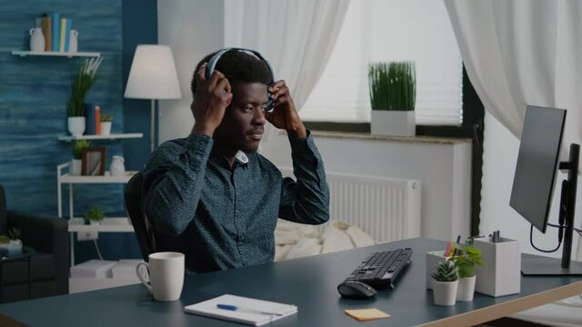 Black Freelancer Of African Ethnicity Putting Headphones On While Working From Home, Remote Worker Computer User In Home Office Typing On PC Listening Music. Concentrated Guy Enjoying Music