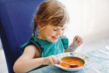 Little preschool girl eating healthy vegetable tomato soup for lunch. Cute happy child taking food...