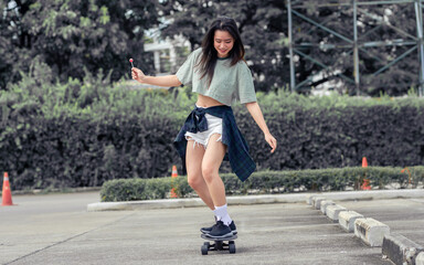 Beautiful happy Asian healthy woman smiling, holding sweet lollipop, riding and playing extreme sportive skateboard as outdoor activity with happiness, relaxation during holidays in summer vacation.