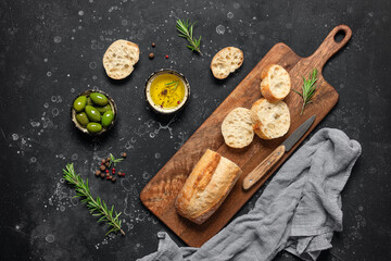 Fresh Italian ciabatta bread in slices with olive oil, olives and rosemary on a black stone background. Top view, flat lay