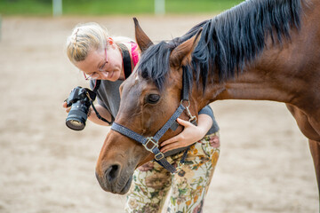 woman with a camera is stroking brown horse's head, girl and horse in the paddock, photographer...