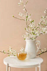 Cup of tasty tea and jug with blooming branches on table near color wall