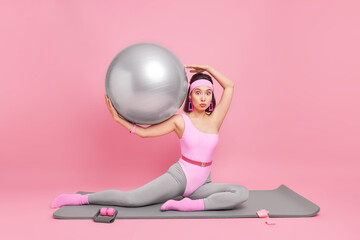 Horizontal shot of surprised Asian woman with fit body does aerobics exercises being fitness instructor works at training centre holds pilates ball dressed in activewear isolated over pink background